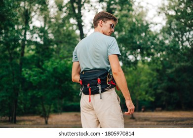 Handsome Young Man In Summer Casual Clothes Posing In The Woods With A Hiking Bag On His Belt, Looking Away. Hipster Man With Waist Bag In The Park.