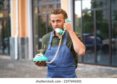 Handsome Young Man Talking On The Vintage, Old Phone Standing Outdoors Wearing Apron. Service Man Having Conversation Using Old Phone. Telephone Communications Concept. 