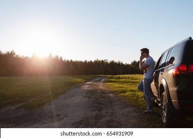Handsome Young Man Talking On Mobile Phone While Leaning On His Car. A Man Watches And Admires In The Direction Of A Beautiful Landscape With A Sunset
