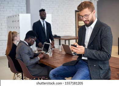 Handsome young man in tuxedo and eyeglasses using smartphone in workplace while his business colleagues having conversation in background - Shutterstock ID 1557181205