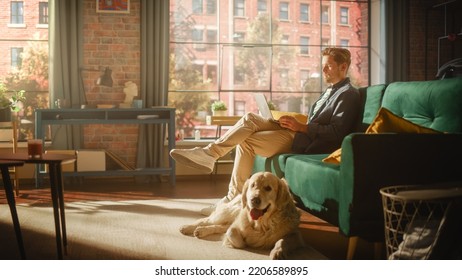 Handsome Young Man Using Laptop, Working From Home Living Room With Big Windows. Male Sitting On Couch, Using Computer, Golden Retriever Dog Sitting Next To Him And Wanting To Play Or Go For A Walk.