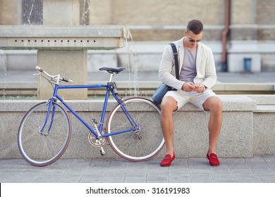 Handsome young man using mobile phone while sitting near the fountain with his bicycle beside him - Powered by Shutterstock