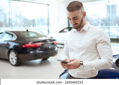 Handsome Young Man Using Smart Phone At Car Dealership, Copy Space. Man Texting On His Phone, Leaning On A Car At Auto Salon