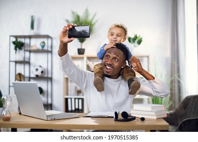 Handsome young man using smartphone for taking selfie with his cute baby son. Happy father sitting at table with modern laptop and documents.