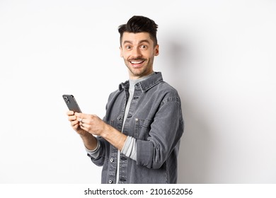 Handsome Young Man Using Smartphone And Looking At Camera. Caucasian Guy Chatting On Phone With Happy Face, Standing On White Background