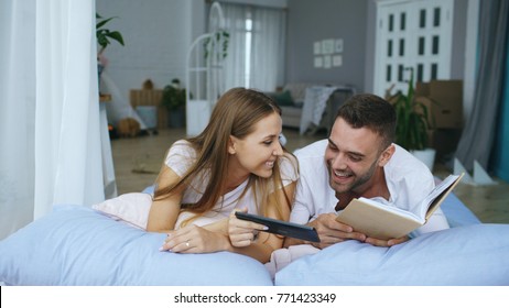 Handsome young man using tablet computer show photos on tablet computer to his girfriend reading book and lying in bed at home