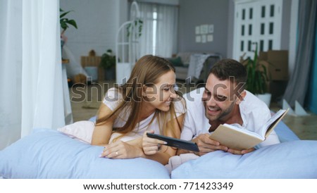 Handsome young man using tablet computer show photos on tablet computer to his girfriend reading book and lying in bed at home