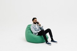 Handsome Young Man Wearing A Casual Outfit, Sitting On A Bean Bag, Holding His Phone Talking To Someone, Looking Away, Isolated On White Background