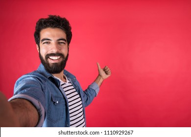 Handsome young man wearing a casual outfit taking a selfie by his phone, raising his thumb up, standing on a red background. - Powered by Shutterstock