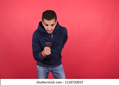 Handsome Young Man Wearing A Casual Outfit, Holding A Magnifying Glasses And Looking Down Searching For Something Missing, Standing On A Red Background.