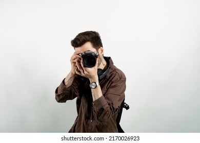Handsome young man wearing casual clothes posing isolated over white background taking images with dslr camera. Photographer covering his face with the camera.