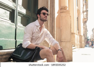 Handsome Young Man Wearing Eyeglasses With Sling Bag Sitting In Modern City Looking Away