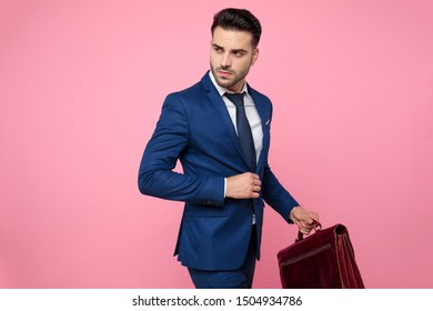 Handsome Young Man Wearing Navy Blue Suit, Holding Suitcase, Fixing Coat And Looking To Side, Standing On Pink Background In Studio