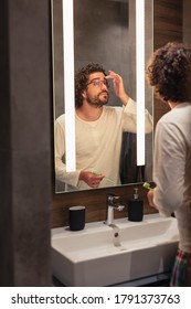 Handsome Young Man Wearing Pajamas Standing In Front Of Mirror In Bathroom, Performing Personal Hygiene Routine At Night Before Sleeping