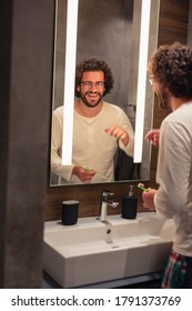 Handsome Young Man Wearing Pajamas Standing In Front Of Mirror In Bathroom, Brushing Teeth At Night Before Sleeping