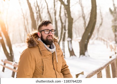 Handsome Young Man Wearing A Warm Winter Clothes, Leaning Against A Wooden Fence And Enjoying A Snowy Winter Day In The Mountains