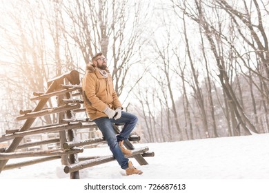Handsome young man wearing a warm winter clothes, leaning against a wooden fence and enjoying a snowy winter day