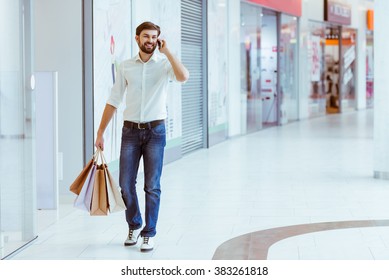 Handsome Young Man In White Shirt Holding Shopping Bags, Talking On A Mobile Phone And Smiling While Walking In Mall