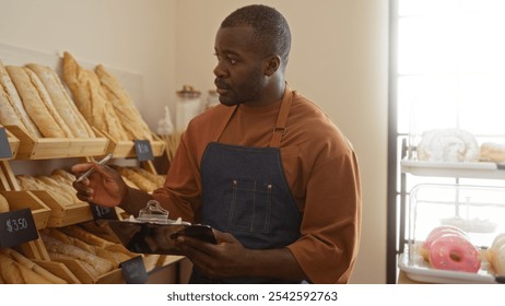 Handsome young man working in a bakery holding a clipboard and pen surrounded by fresh bread loaves and pastries indoors - Powered by Shutterstock