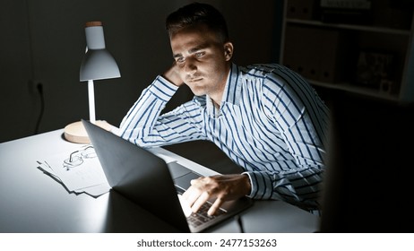 Handsome young man working late on laptop in a dimly lit modern office, portraying dedication. - Powered by Shutterstock