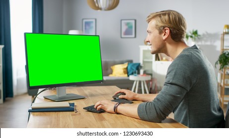 Handsome Young Man Works On A Green Mock-up Screen Personal Computer While Sitting At His Desk In The Cozy Apartment.