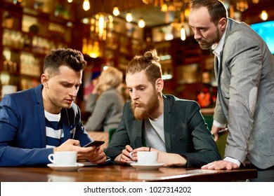 Handsome young manager using smartphone while having informal working meeting with colleagues, interior of modern cafe on background