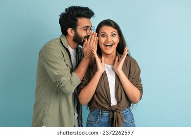 Handsome young middle eastern guy sharing secret or whispering gossips into his girlfriend's ear, excited pretty brunette indian woman gesturing, isolated on blue studio background, copy space