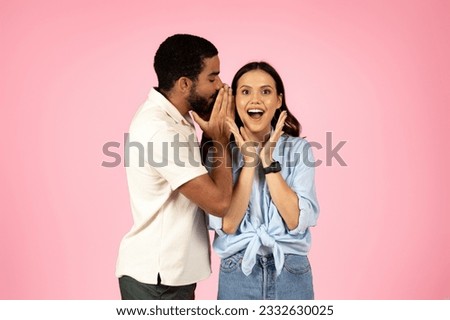 Handsome young middle eastern guy sharing secret or whispering gossips into his girlfriend's ear, excited pretty brunette woman gesturing, isolated on pink studio background, copy space