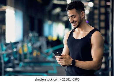 Handsome Young Middle Eastern Man Using Smartphone During Training In Sport Club, Smiling Arab Male Athlete Messaging On Cellphone Or Browsing New Fitness App While Spending Time At Gym, Copy Space - Powered by Shutterstock