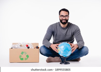 Handsome young moroccan man in casual attire sitting with a box full of plastic bottles for recycling isolated over white background. Save the planet concept