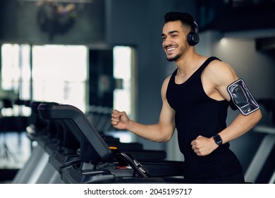 Handsome Young Muscular Arab Man In Wireless Headphones Jogging On Treadmill At Gym, Sporty Motivated Middle Eastern Guy Listening Music While Running, Enjoying Making Sports, Copy Space