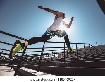 Handsome Young Muscular Man In Modern Sport Clothing Run Up The Stairs Outdoor At Bright Sunny Day. Wide Angle Photo Of A Jogging Man. Sport Lifestyle