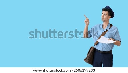 Handsome young postman pointing at something on light blue background with space for text