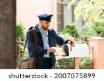 Handsome young postman putting letter in mail box outdoors