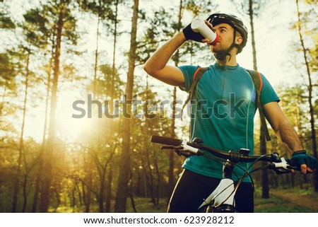 Handsome young professional cyclist dressed in cycling clothing and protective helmet feeling free and happy, drinking water from the bottle, enjoying bicycle in warm spring. Recreation of sportsman.