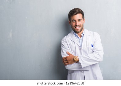 Handsome young professional man over grey grunge wall wearing white coat happy face smiling with crossed arms looking at the camera. Positive person.