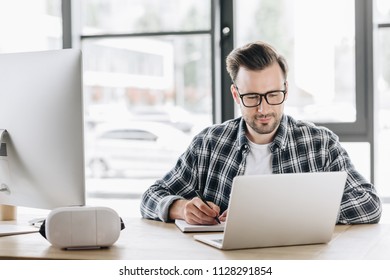 Handsome Young Programmer In Eyeglasses Taking Notes While Working With Laptop And Desktop Computer