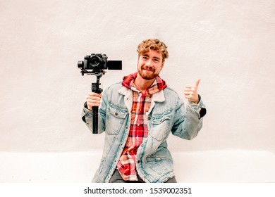 A Handsome Young Red Head Guy Smiling And Wearing Denim Holding A Camera With Steadicam Stabilizer And Showing Thumb Up