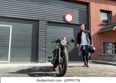 Handsome young rider guy in black biker jacket and jeans go to his classic style cafe racer motorcycle industrial gates as background. Bike custom made in vintage garage. Brutal fun urban lifestyle. - Powered by Shutterstock