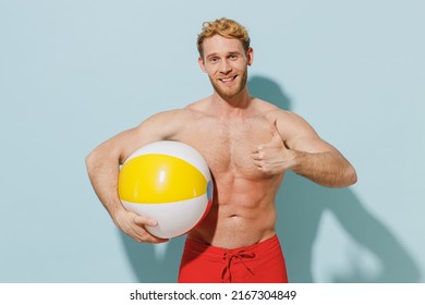 Handsome young sexy smiling happy man in red shorts swimsuit relax near hotel pool hold inflatable rubber ball show thumb up gesture isolated on blue background Summer vacation sea sun tan concept.