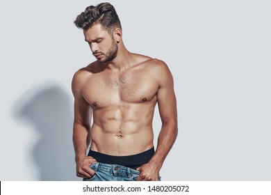 Handsome young shirtless man taking off his jeans while standing against grey background