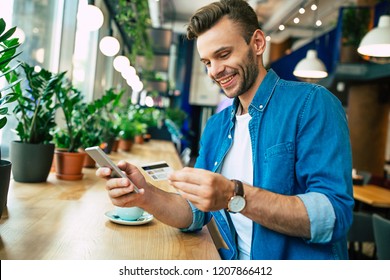 Handsome young smiling man with credit card and phone in hands doing purchases during online shopping 