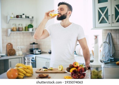 Handsome young sporty smiling man in the kitchen is preparing vegan healthy fruits salad and smoothie in a good mood