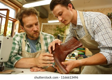 Handsome young tailor showing brown leather boot to senior tailor, inspecting product for correct sewing. Two men wearing in aprons, chatting in room with sewing machines at factory. Teamwork concept. - Powered by Shutterstock