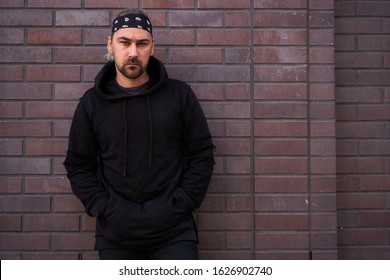 Handsome Young Unshaven Man Black Hoodie And Bandana Stands Backyard Near Brick Walls Summer Day. Bearded Hipster Standing Outdoor. Fashionable Young Adult Caucasian Guy Modern Urban Clothes