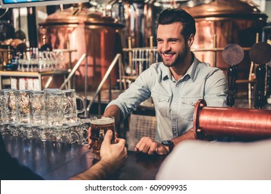 Handsome young waiter is giving beer to clients and smiling while working in pub