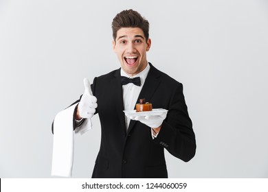 Handsome Young Waiter In Tuxedo With Bowtie Holding Plate With Cake Isolated Over White Background, Thumbs P