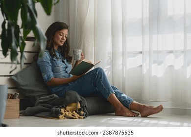 Handsome young woman reading a book, drinking coffee at home sitting cozy by the window - Powered by Shutterstock