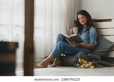 Handsome young woman reading a book, drinking coffee at home sitting cozy by the window - Powered by Shutterstock