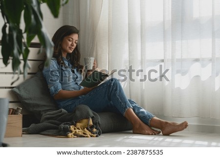 Handsome young woman reading a book, drinking coffee at home sitting cozy by the window